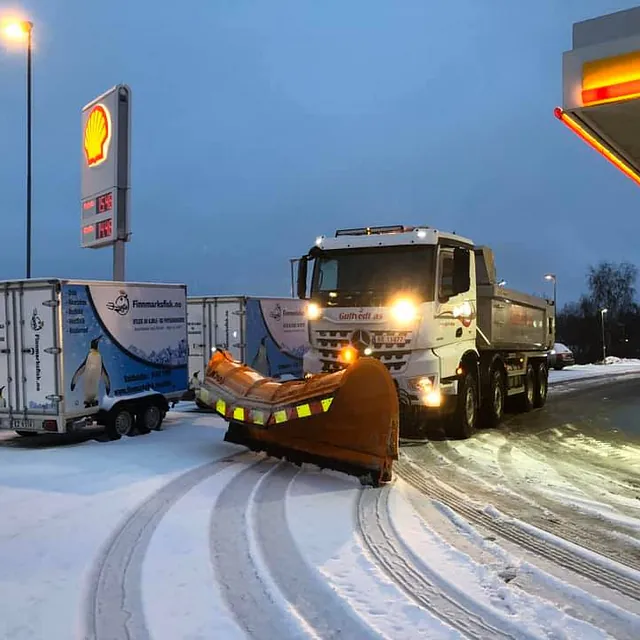 Snøbrøytebil kjører forbi Shell-stasjon i vinterlys, med snøplog og henger på parkeringsplass.