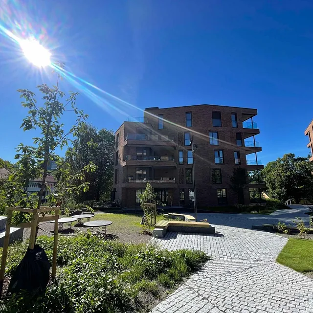 Solfylt tun mellom moderne mursteinsbygg med brostein, trær og benker i grøntområde.