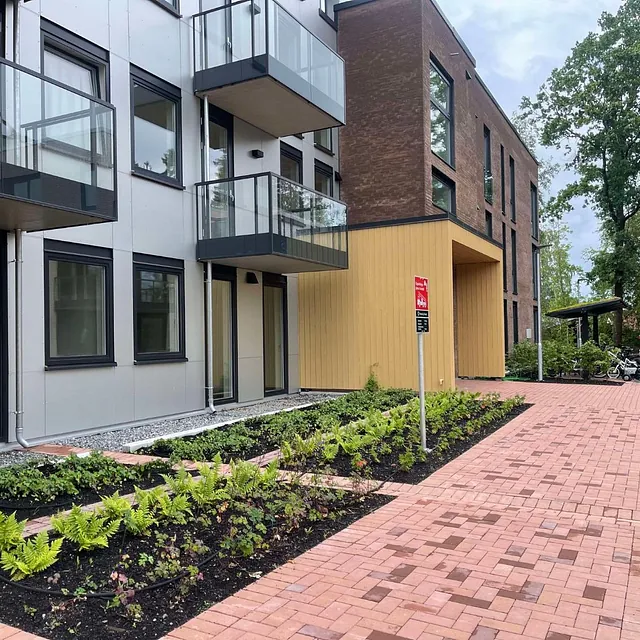 Inngangsparti til moderne leilighetsbygg med belegningsstein, sykkelparkering og beplantning.