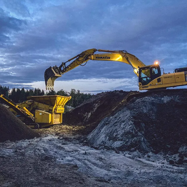 Gravemaskin arbeider i skumringen og fyller en stor sorteringsmaskin med jord og stein.