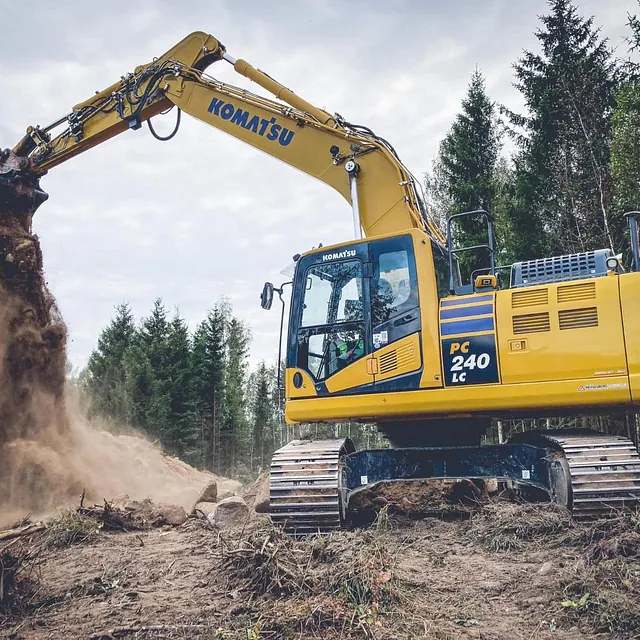 Gravemaskin fra Komatsu tipper jord mens den jobber på et skogsområde med ujevnt terreng.